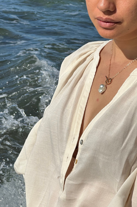 A close-up lifestyle shot of a woman wearing a cream-colored linen button-up shirt and a gold necklace with a Ursa Major pearl pendant, set against a background of ocean water.